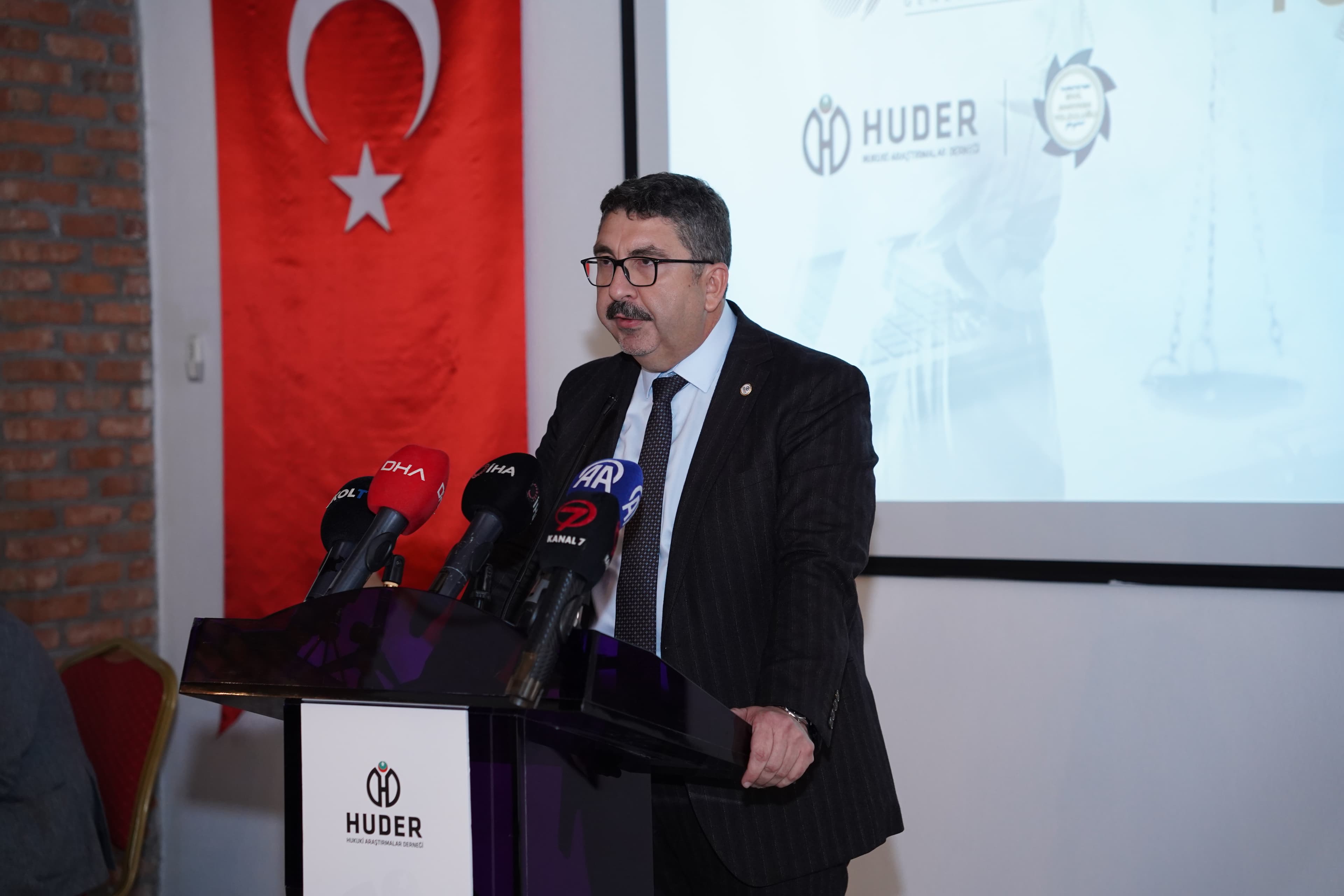 President Hasan Oymak speaking at the HUDER podium in front of the press microphones; behind him, the Turkish flag.